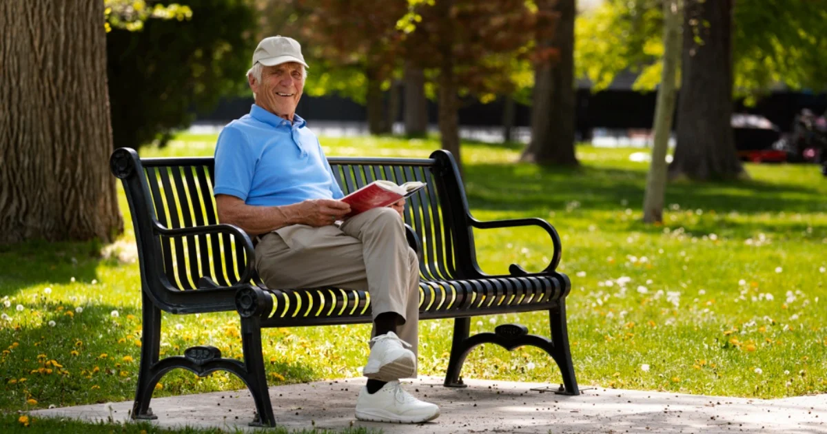  Smiling senior man reading a book while sitting on a park bench in a sunny, green outdoor setting.